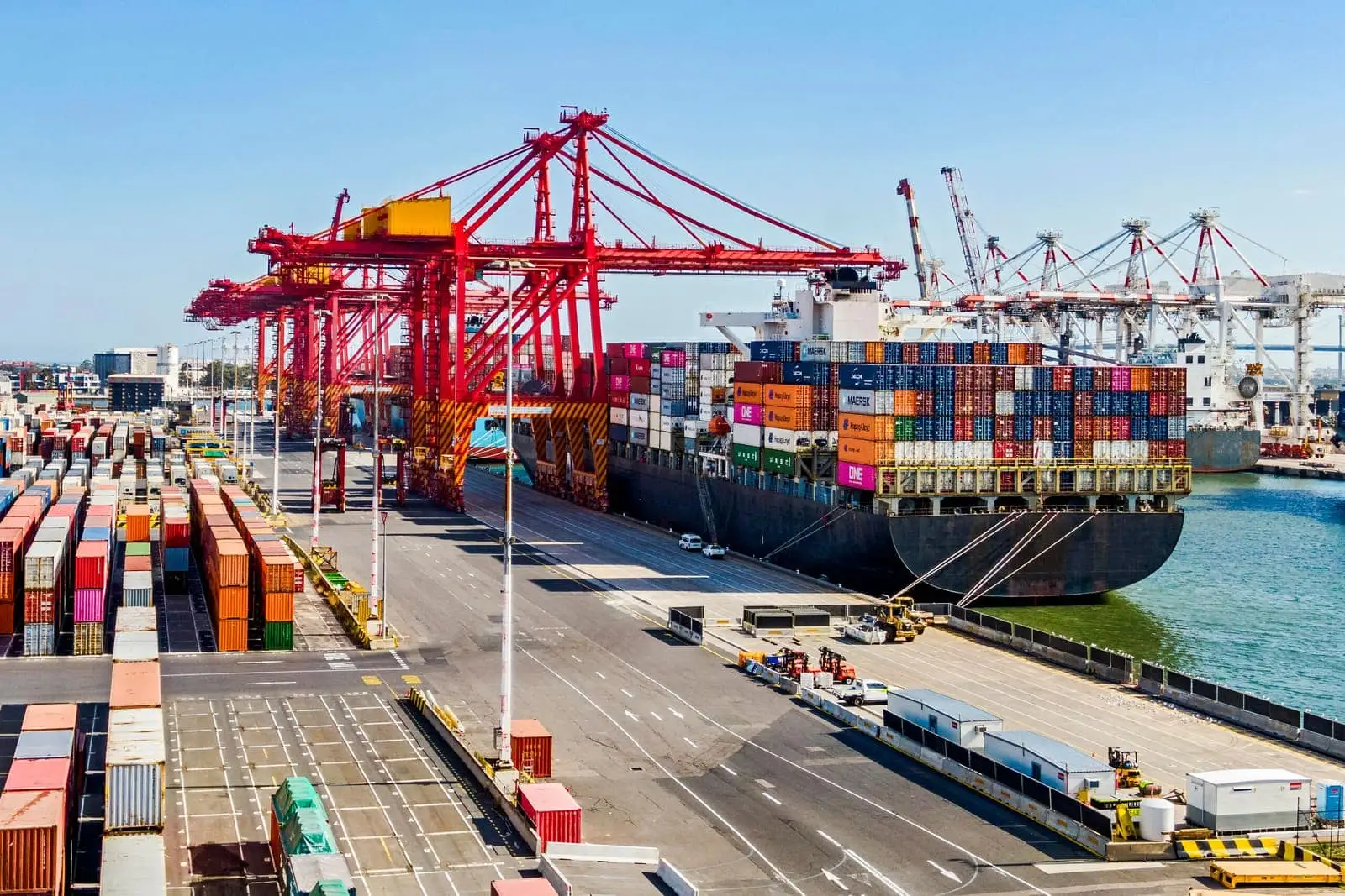 Container ship loading at a busy port terminal with red gantry cranes