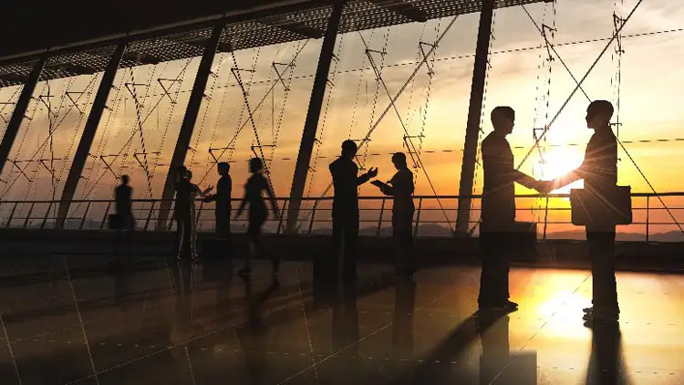 Two business people shaking hands at sunset while others network nearby