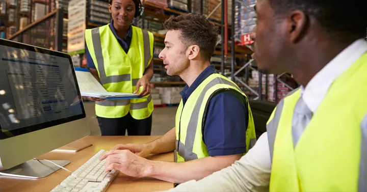 Three warehouse workers in high-visibility vests reviewing inventory records on a desktop computer among storage racks.