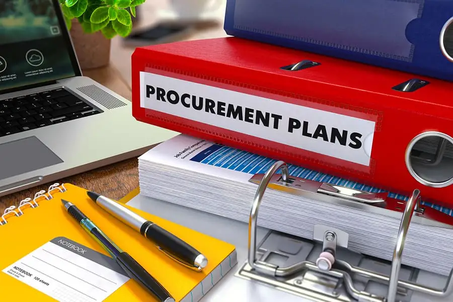 A red binder labeled Procurement Plans stacked on a desk with a laptop and notebook